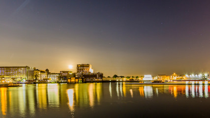 Obraz premium Night view of buildings on the shore of new Maas in business city of Rotterdam, yellow lights reflected on water surface, calm night in South Holland in the Netherlands. Long exposure photography
