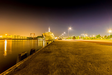 Fototapeta na wymiar Night view of the port of Rotterdam with a cruise ship anchored on the quay, empty street with illuminated lamp posts and trees in the background, quiet night with a clear sky in the Netherlands