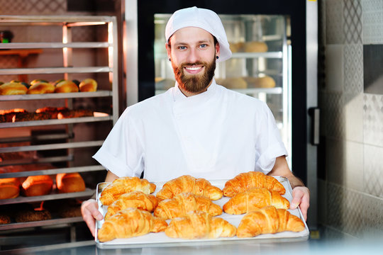A young handsome male baker holds a tray with French croissants in front of a bakery and smiles.