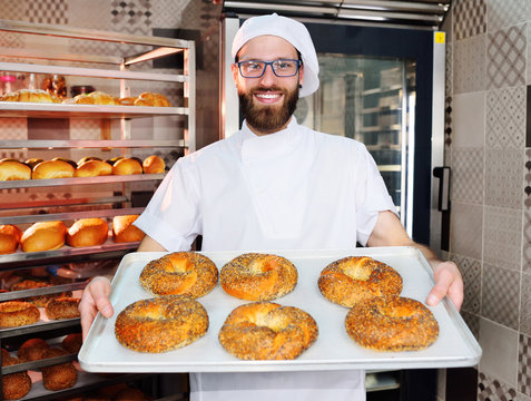attractive Baker in white uniform holding a tray with freshly baked bagels with sesame and poppy seeds on the background of a bread factory or bakery