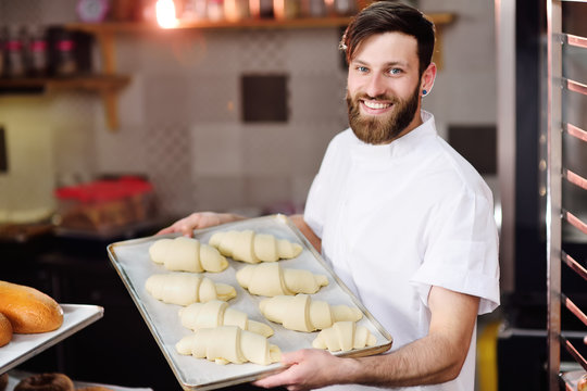 a handsome Baker with a beard prepares croissants for baking and smiles at the bakery background