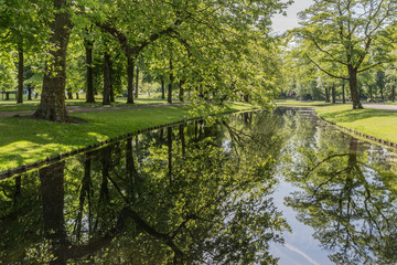 Stream with calm water and mirror reflection on the water surface of the surrounding lush green trees, calm sunny day in a public park in the city of Rotterdam, South Holland, Netherlands