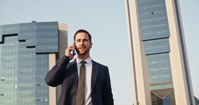 Multiethnic businessman walking down street between scyscrapers with briefcase in hand, talking on phone, and smiling closeup 4k