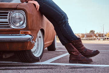 Cropped photo of male in jeans and boots leaning on retro car in the city parking.