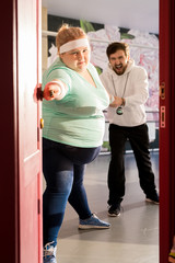 Fototapeta premium Full length portrait of fat woman trying to escape training in fitness club with coach struggling to hold her