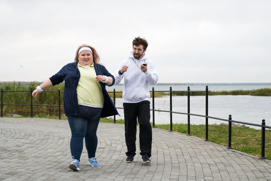 Full length portrait of overweight woman running outdoors with personal trainer motivating her, copy space