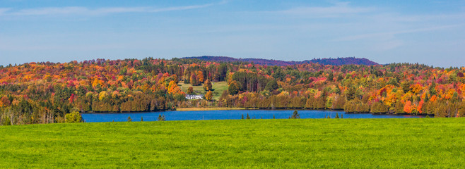 panorama banner of white house in the country  with blue pond, green fields and  trees  dressed in bright autumn colors of fall foliage 
