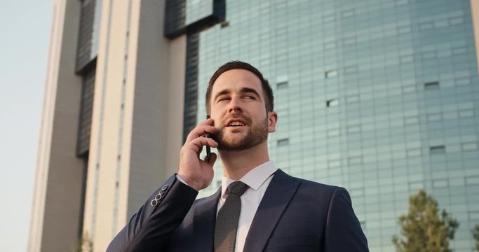 Multiethnic businessman standing in street between scyscrapers with briefcase in hand, talking on phone, and smiling closeup 4k