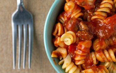 Top close view of a bowl of rotini pasta in a sausage and tomato sauce with a fork to the side on a brown tablecloth.
