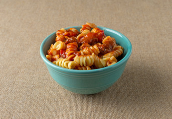 A bowl of rotini pasta in a sausage and tomato sauce on a brown tablecloth.