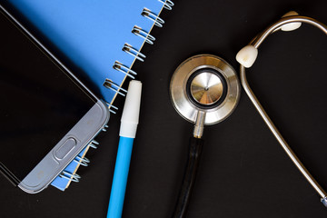 Medical conceptual image with the view of stethoscope, blue pen, book and smartphone on the black background.
