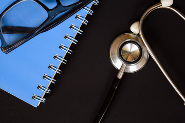 Medical conceptual image with the view of stethoscope, eyeglasses and blue book on the black background.
