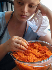 Young girl in apron and a cook's cap prepares fresh carrot salad in kitchen. Woman cook rubs carrots through grater, adds garlic, salt and mayonnaise