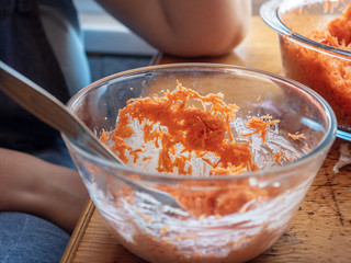 Young girl in apron and a cook's cap prepares fresh carrot salad in kitchen. Woman cook rubs carrots through grater, adds garlic, salt and mayonnaise