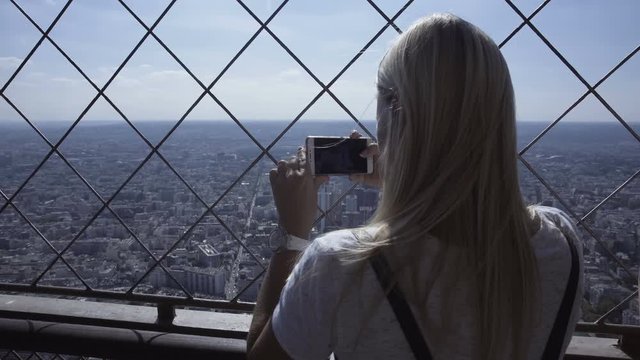 Back view of blond woman on Eiffel tower taking photo of city panorama with smartphone in sunlight