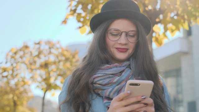 Close up portrait of texting woman on a smartphone device. Beautiful young woman wearing hat and glasses using modern smart phone typing text message on cellphone outside