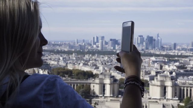 Back view of blond woman using phone and taking photo of new cityscape from Eiffel Tower in sunlight