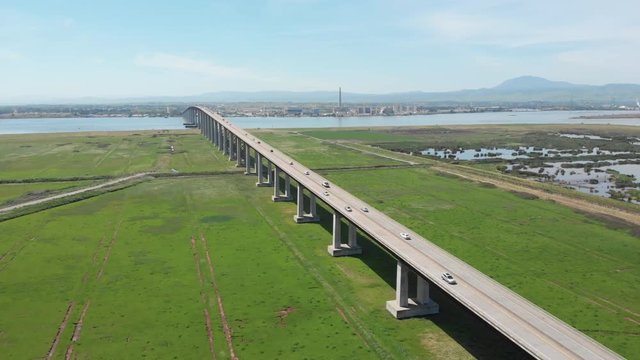 AERIAL: Countless cars driving to and from a large industrial town in the picturesque sunny rural part of California. Flying along spectacular concrete bridge leading freeway traffic across the river.