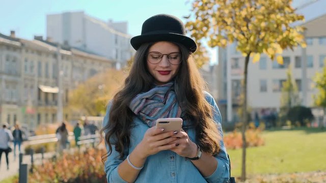 Close up portrait of smiling woman holding smartphone looking to camera. Young girl looking at camera at autumn city background.