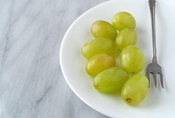 Side view of a serving of green grapes with a fork to the side on a white plate atop a marble counter top.