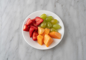 Overhead view of strawberries grapes and cut cantaloupe on a white plate atop a gray marble counter top.