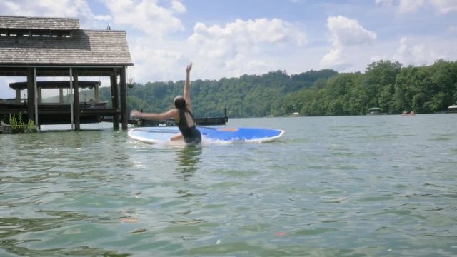 Wide Shot Of A Young Woman Falling Off A Stand Up Paddle Board In Slow Motion. Standing Upright The Millennial Girl Paddles Her Floating Vessel Forward Then Clumsily Falls Into The Water. 