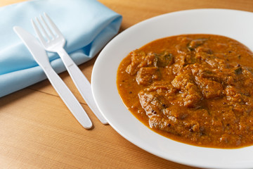 A meal of eggplant curry on a white plate with a plastic fork and knife and a blue napkin to the side atop a wood table top.