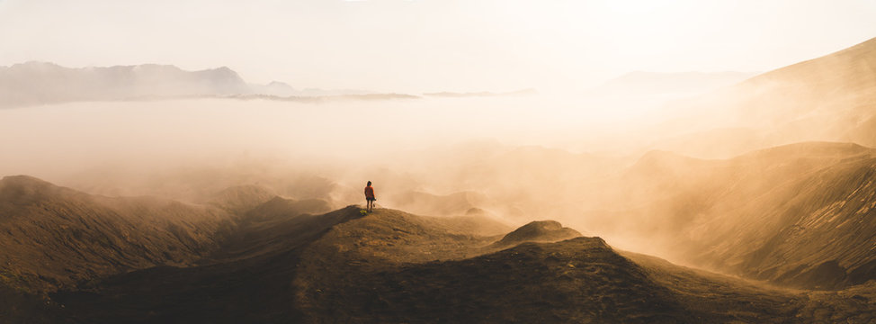 Panoramic of a young woman standing at the edge of a volcano looking into the distance.