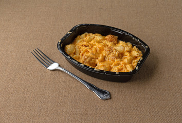 A spicy chicken nuggets mixed in with macaroni and cheese TV dinner in a black tray with a fork to the side on a coarse brown tablecloth.