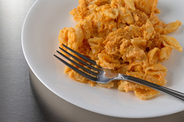 Close view of a serving of spicy chicken nuggets mixed in with macaroni and cheese on a white plate and a fork atop a stainless steel counter top.