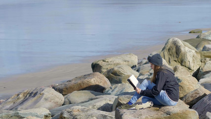 Lonely girl reads a book on the rocks at the waterfront