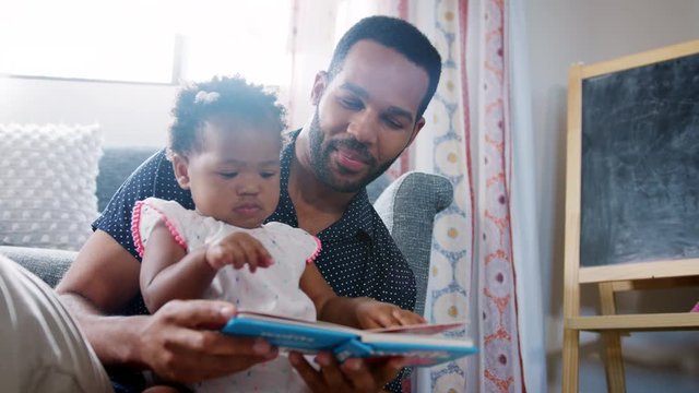 Father Sitting On Floor And Reading Book With Baby Daughter At Home