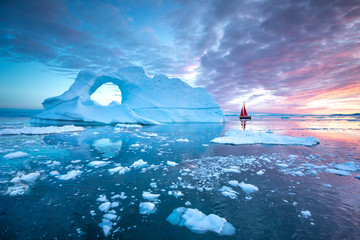 Little red sailboat cruising among floating icebergs in Disko Bay glacier during midnight sun...