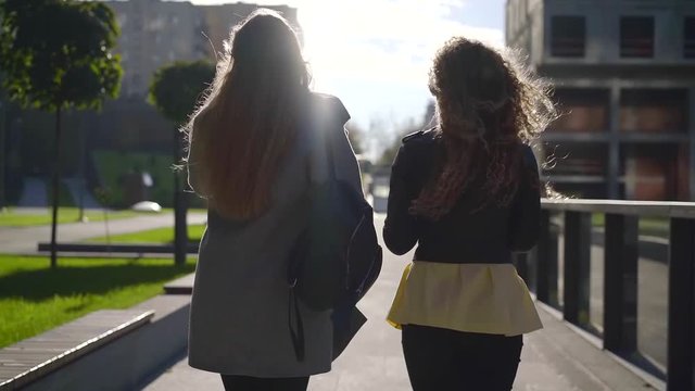 rear view of two young slim women are walking on alleys in park, against sun in autumn day, students