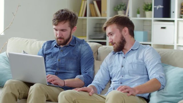 Two young Caucasian twin brothers sitting on sofa in living room at home and using laptop computer