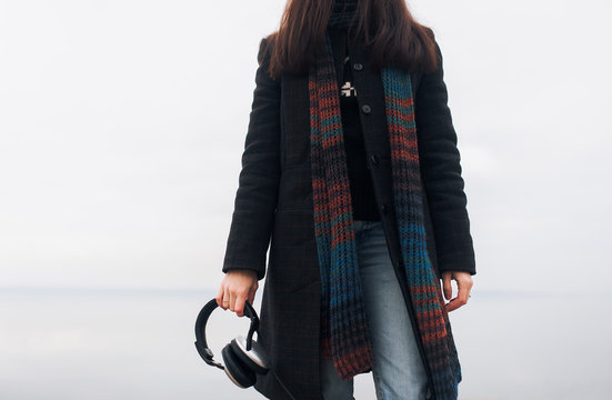 The girl in the coat holds big headphones in her hands. Listen to music on a walk in earphones.