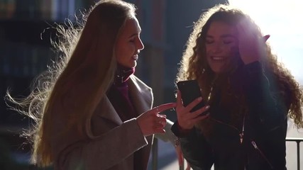charming young sisters are having fun outside in windy autumn day, watching pictures on mobile phone - Powered by Adobe