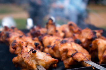 Skewers with roasted chiken meat on the hot grill closeup with two blurred human silhouettes in the background