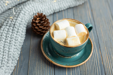 Cup of coffee, marshmallow, warm knitted sweater on wooden background. Warm lights. Cozy winter morning. Lifestyle concept. Selective focus.