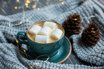 Cup of coffee, marshmallow, warm knitted sweater on wooden background. Warm lights. Cozy winter morning. Lifestyle concept. Selective focus.