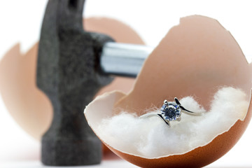 The egg shell is broken by a hammer and fake diamond ring in the egg shell on white background....