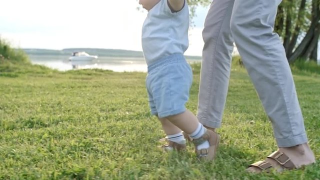 Tracking of unrecognizable woman holding hands of toddler boy walking on green grass in park on summer day; lake in background