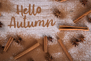 Hello, Autumn. The phrase is written on a wooden table with ground cinnamon among cinnamon sticks and badyan.  Top view, flat lay