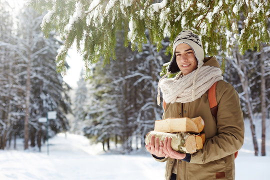 Waist up portrait of handsome young man posing in winter forest holding logs and smiling at camera cheerfully, copy space