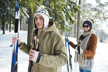 Waist up portrait of happy young couple skiing in winter forest, focus on man in front smiling at camera