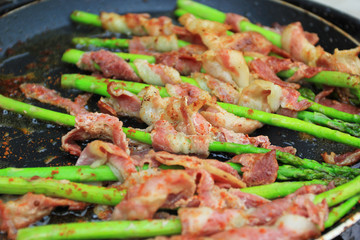 Raw bacon wrapped green asparagus in frying pan on dark background