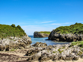 Beautiful and lovely beach Playa de Buelna with blue ocean water, rocky cliffs covered with green vegetation and blue sky in background. Sea landscape of the coast of Bay of Biscay, Asturias, Spain