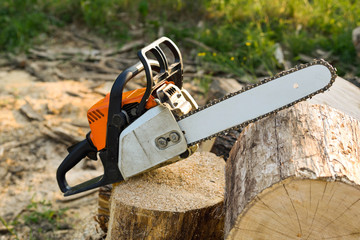 Fototapeta premium Chainsaw that stands on a heap of firewood in the yard on a beautiful background of green grass and forest.