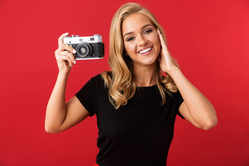 Portrait of young blond woman 20s smiling and photographing on retro camera, isolated over red background in studio