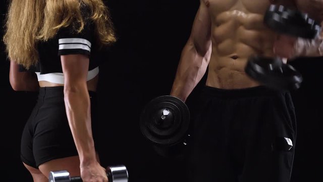 Athletic man and woman flexes their hands with dumbbells, training their biceps on a black background in studio